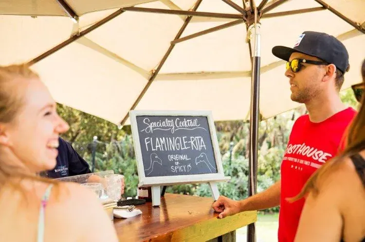 People at an outdoor bar ordering flamingo-themed drinks under a white umbrella
