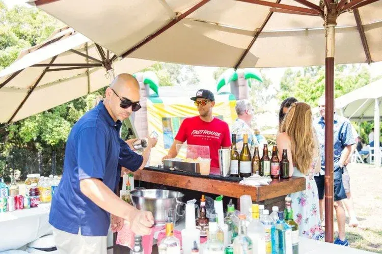 People serving drinks at an outdoor bar under a white canopy, with bottles and cups on display