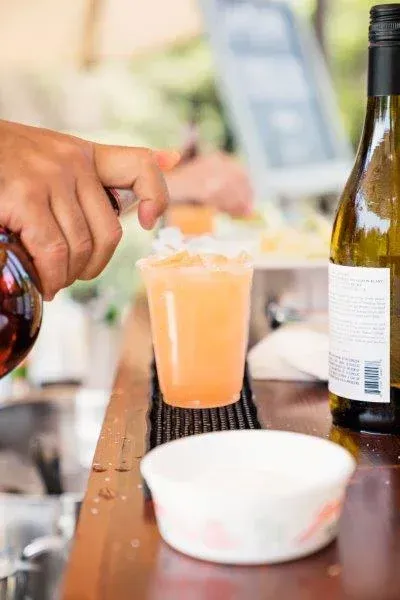 Hands pouring a drink into an orange cocktail on a bar counter with a wine bottle nearby