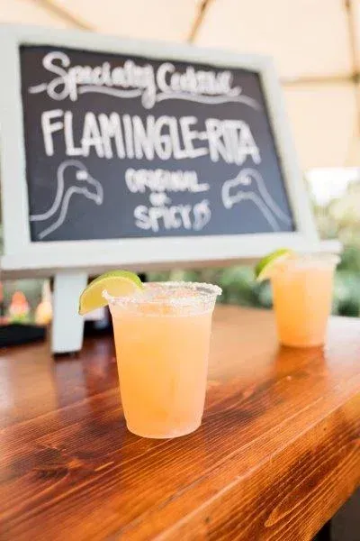Two orange cocktails on a wooden bar with a chalkboard sign reading “Spaghetti Western Flamingolita”