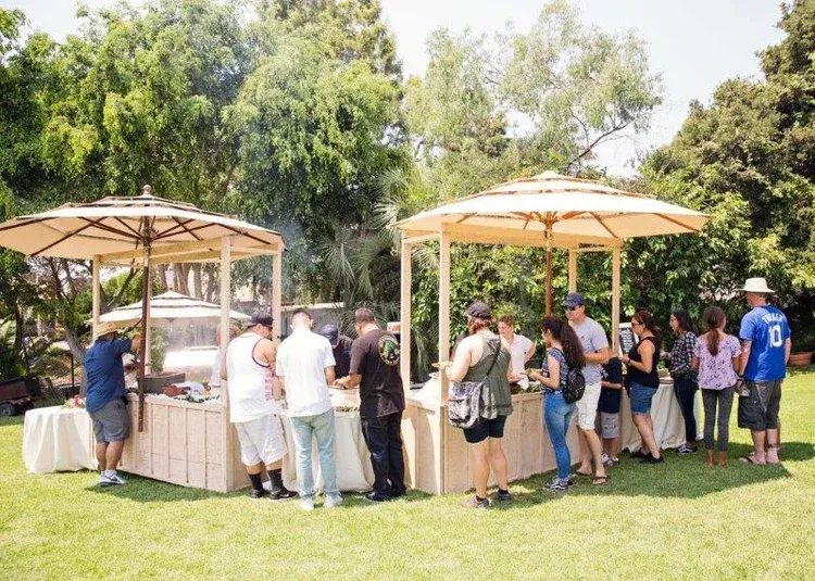 People gathered around outdoor buffet tables under umbrellas in a sunny park.