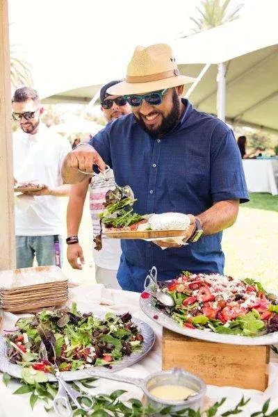 Man serving a salad buffet outdoors under a white tent, with large bowls of fresh greens.