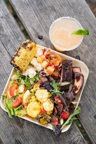 Grilled vegetable salad with corn, tomatoes, greens, and a drink in a takeout container on a wooden table