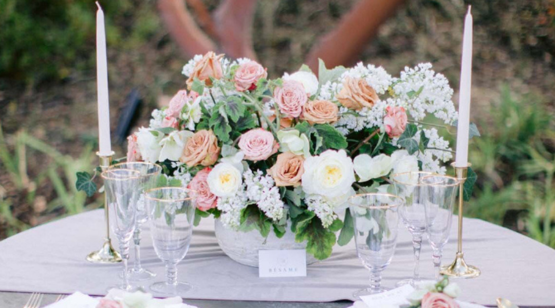 Elegant floral table centerpiece with blush roses, white flowers, candles, and glassware on a round table outdoors