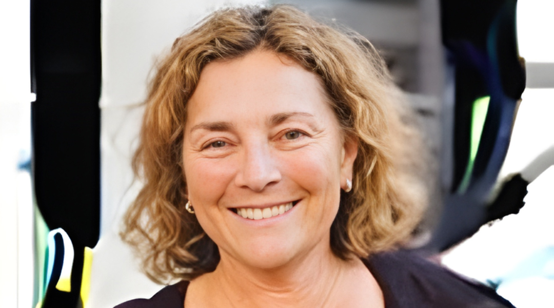 Smiling person with curly blond hair, seated against a bright outdoor background