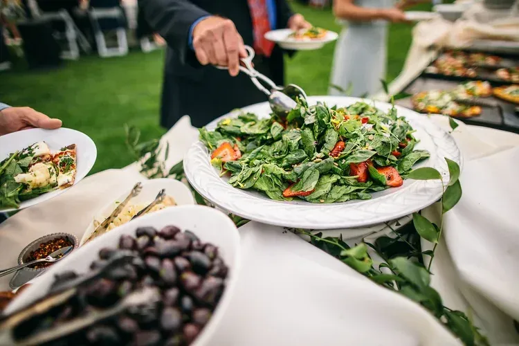 Buffet salad being served at an outdoor event with bowls of greens and olives