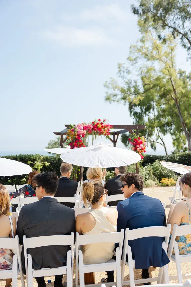 Guests seated outdoors under white umbrellas at a sunny wedding ceremony with floral decorations
