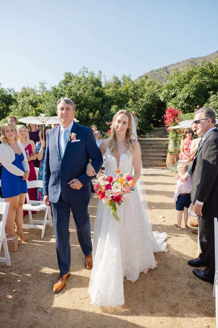 Bride and groom walking outdoors at a sunny wedding ceremony, bride holding a colorful bouquet