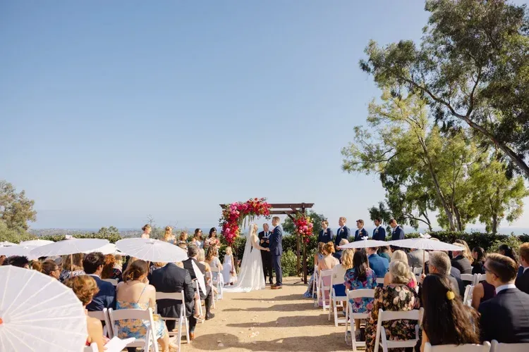 Outdoor wedding ceremony with guests seated along an aisle beneath a floral arch under a clear blue sky