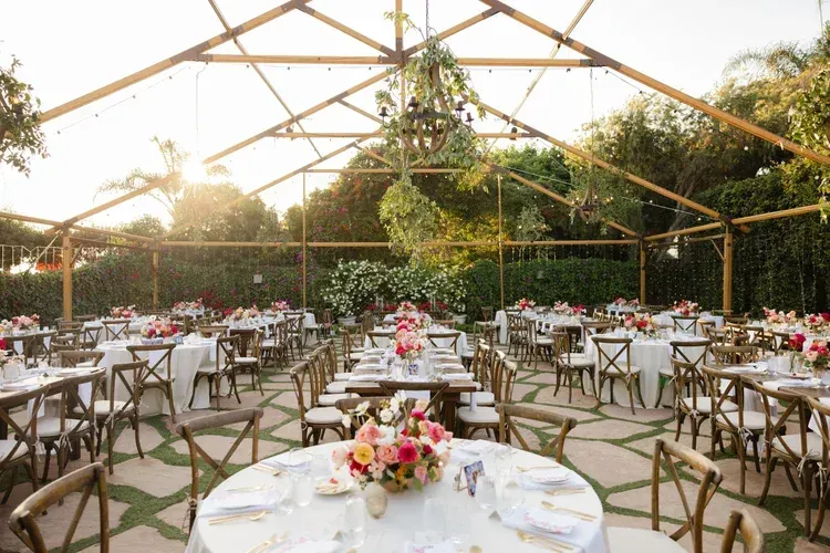 Elegant outdoor wedding reception with round tables, floral centerpieces, and a canopy over a garden setting.
