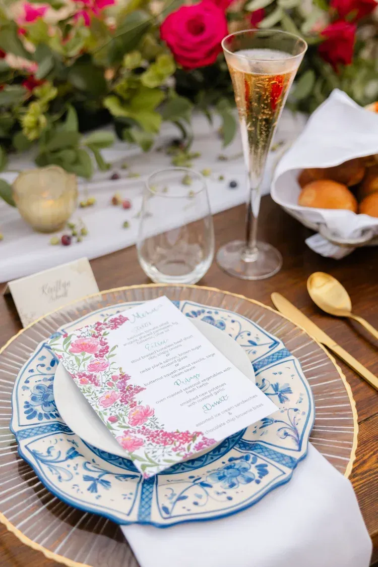 Elegant table setting with floral plates, menu card, champagne flute, and pastries amid pink roses