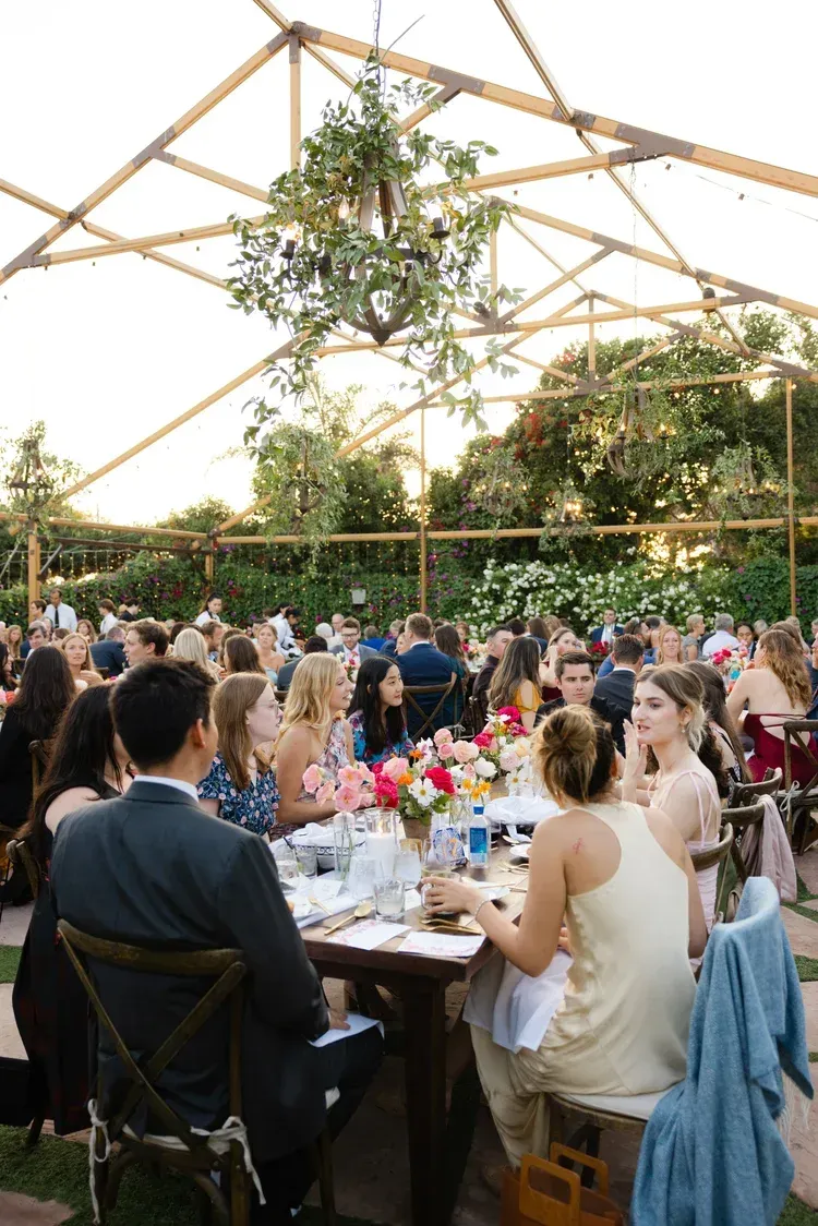 Guests dining at a wedding reception under a wooden pavilion with hanging greenery and string lights.