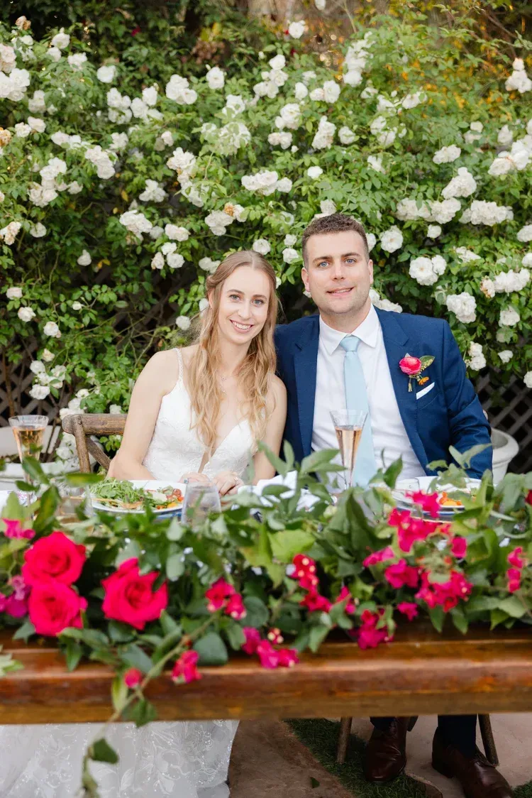 Newlyweds seated at a flower-filled wedding table with white and pink blooms behind them