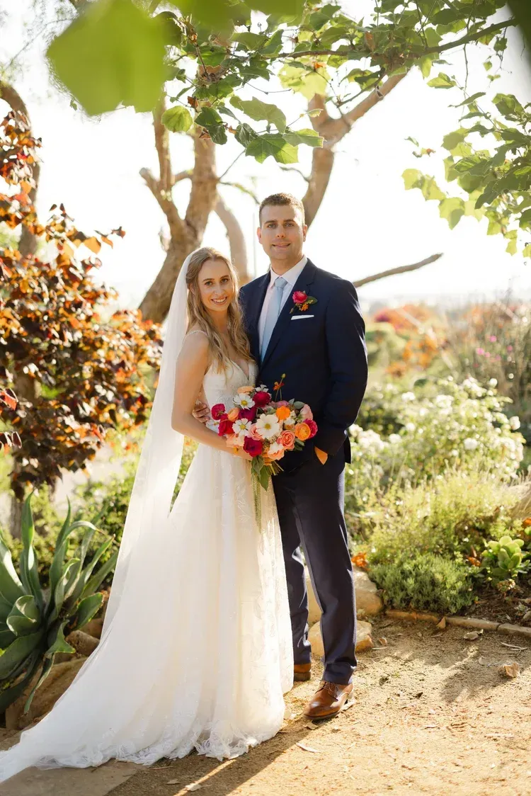 Bride and groom in wedding attire standing outdoors, holding a colorful bouquet under sunlit trees
