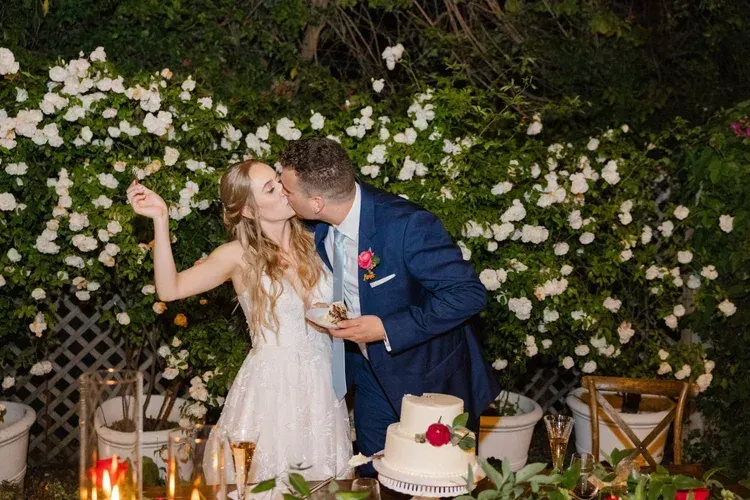 Couple kissing at a decorated outdoor wedding reception beside a cake and flowers