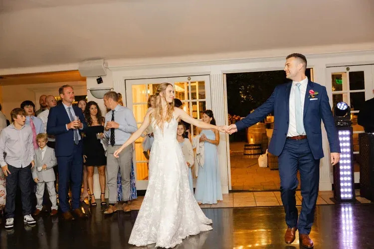 Bride in a white gown and groom in a blue suit walk hand in hand at a wedding reception, guests watching.