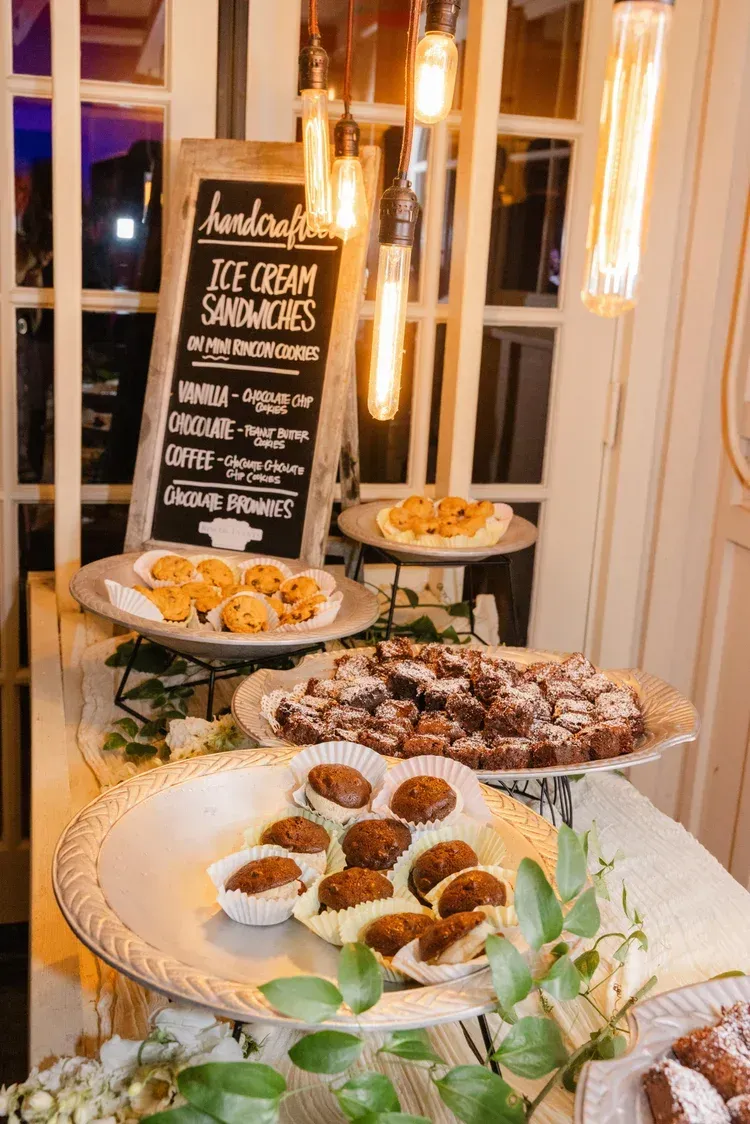 Dessert buffet with pastries and doughnuts on tiered stands beside a chalkboard menu and warm string lights