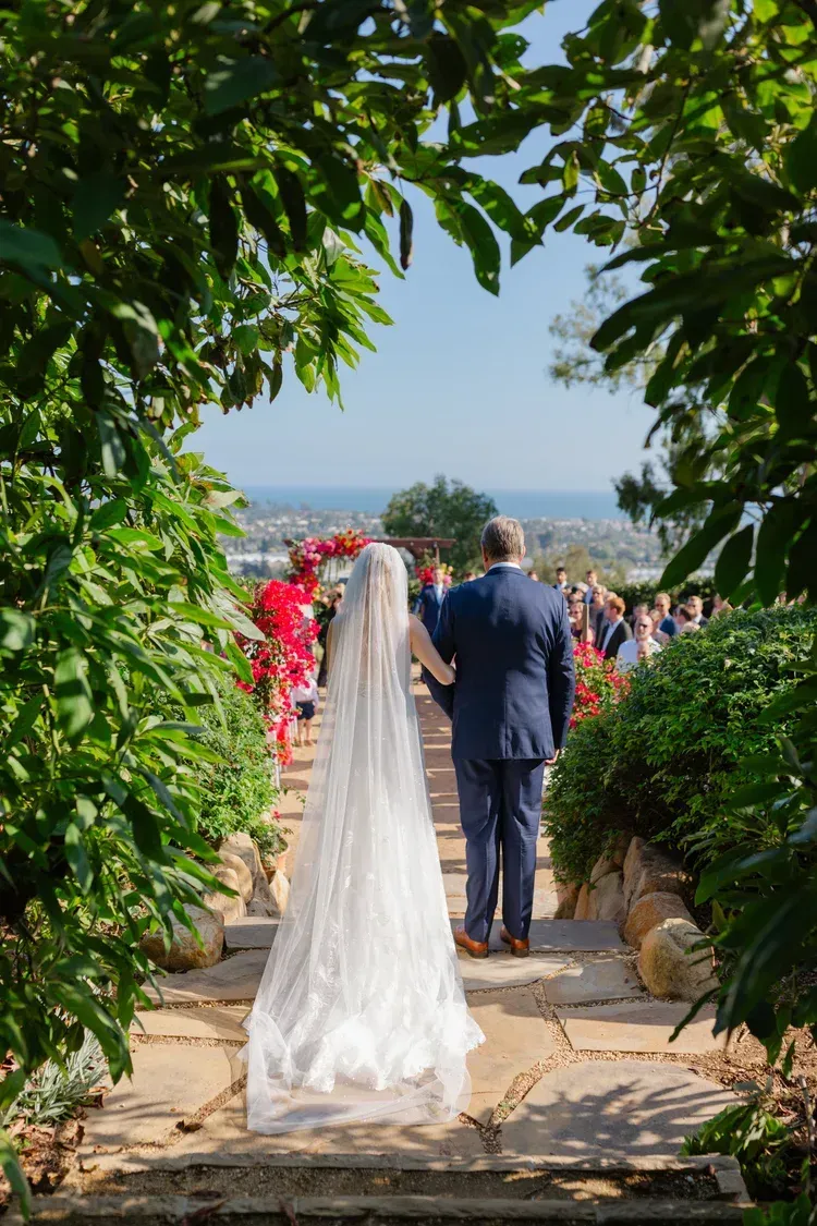 Bride in a white veil and groom in a navy suit walking down a flower-lined aisle outdoors