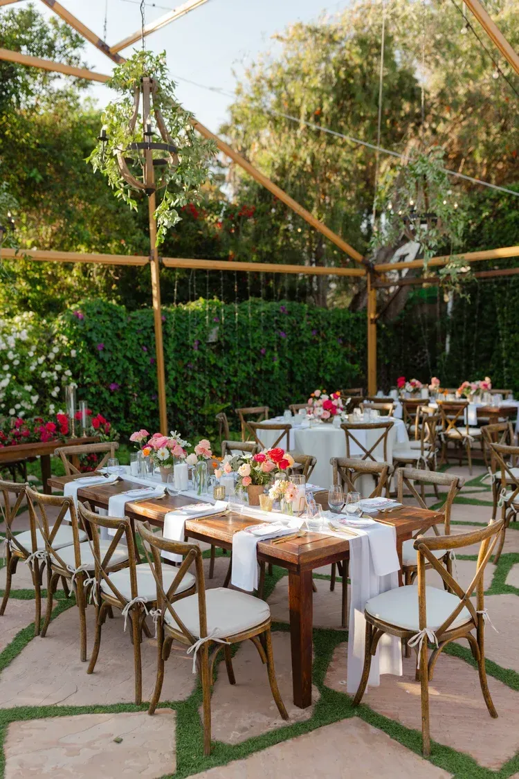 Outdoor event tables set under a rustic canopy with white chairs and pink floral centerpieces