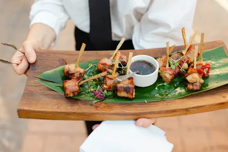 Person holding a wooden platter with grilled skewers and dipping sauce on a banana leaf at a table