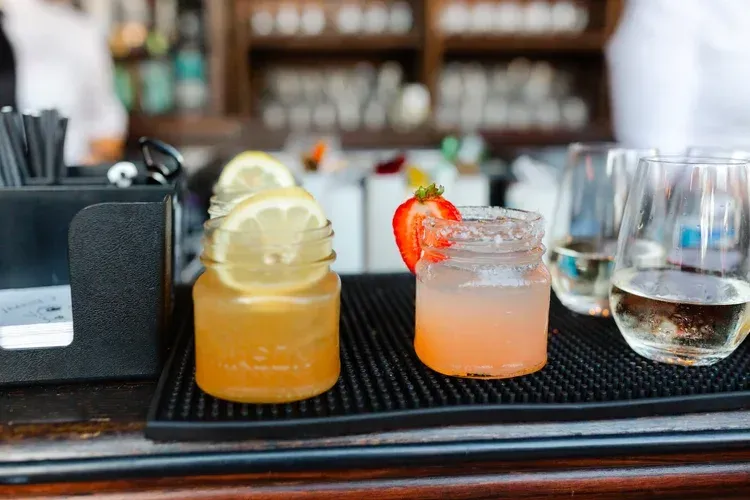 Two cocktails on a bar tray, one orange with lemon slices and one pink with a strawberry garnish.
