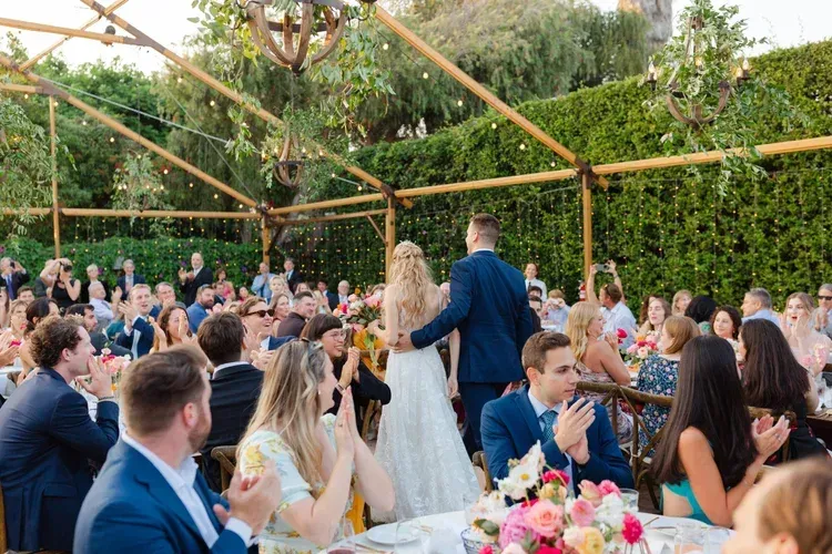 Outdoor wedding reception with guests seated at decorated tables under a wooden pergola