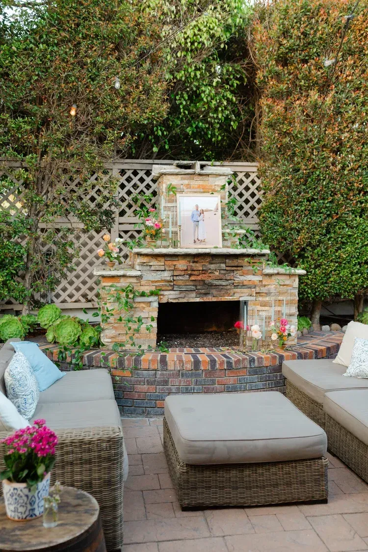 Patio seating area with wicker chairs and ottoman around a brick fireplace framed by greenery