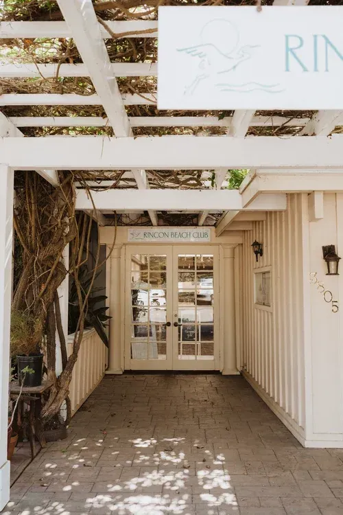 Entrance to a white wooden building with a pergola and glass doors, lit by dappled sunlight.