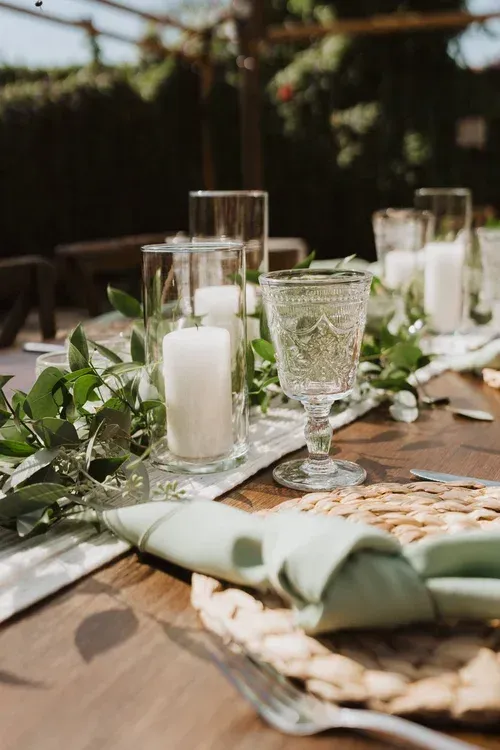 Greenery and glassware on a set outdoor table with white candles and woven placemats