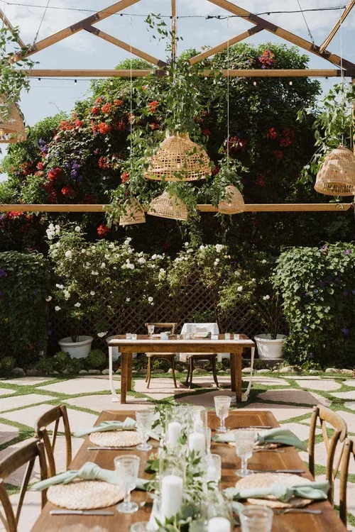 Elegant outdoor dining setup under a glass greenhouse canopy, with lush hanging flowers and wooden tables.