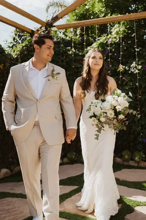 Bride and groom holding hands and bouquet, walking outdoors beneath a wooden arch.