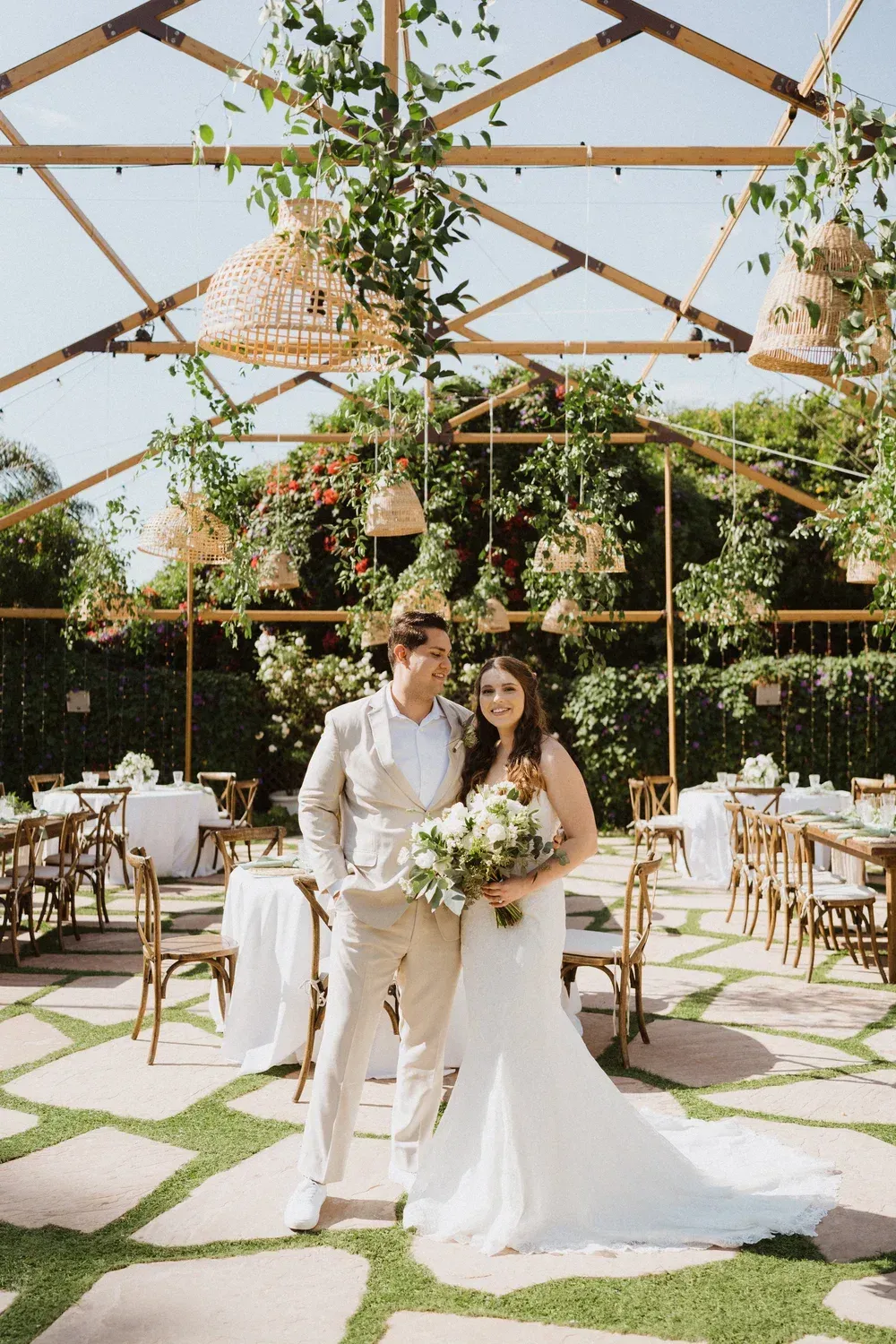 Bride and groom posing in a flower-filled outdoor wedding venue under a wooden canopy