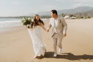 Couple in wedding attire running hand in hand on a sunny beach, bride holding a bouquet.