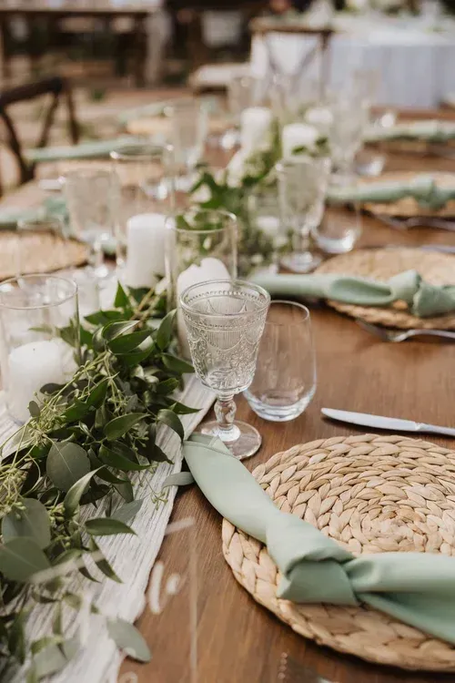Elegant outdoor banquet table with woven placemats, green napkins, candles, and white floral greenery.
