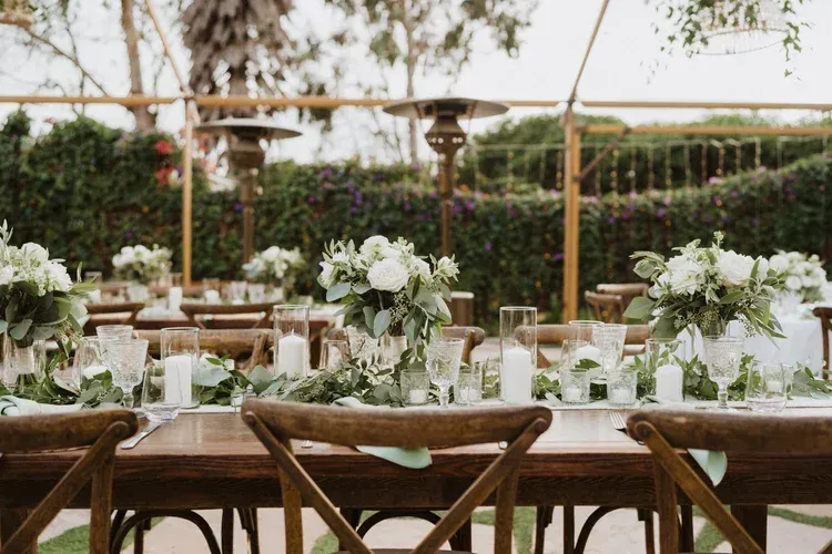 Elegant outdoor wedding reception table with white floral arrangements and wooden chairs under greenery