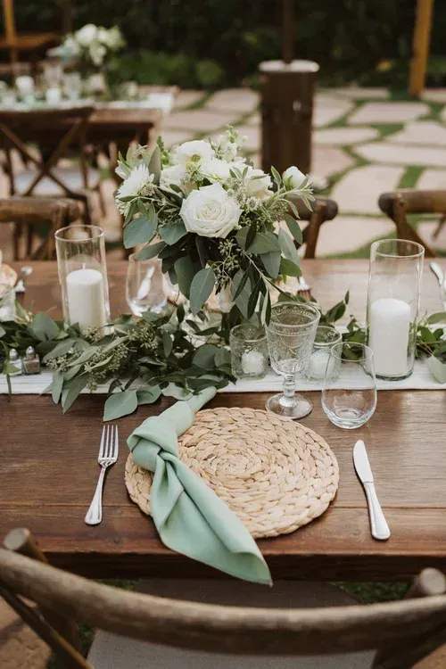 Elegant outdoor table setting with white floral centerpiece, glassware, candles, and sage-green napkin