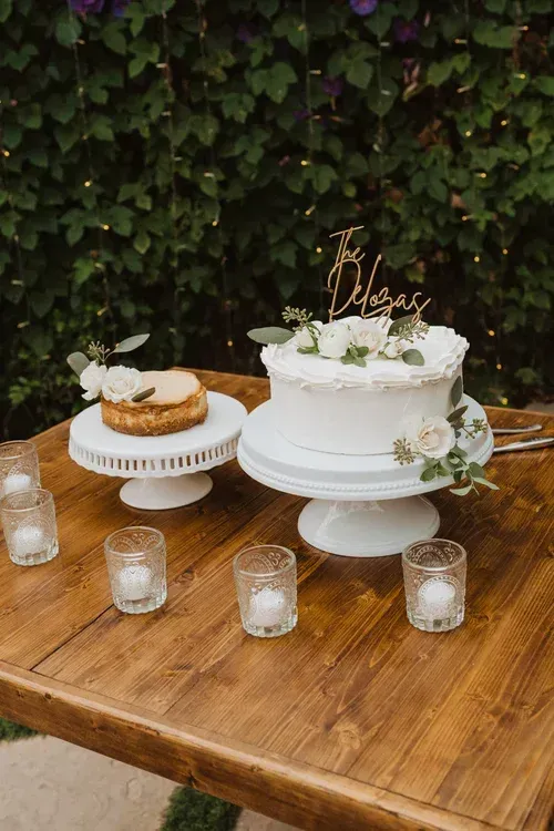 White wedding cake and small dessert on a wooden table with glass votives and greenery backdrop