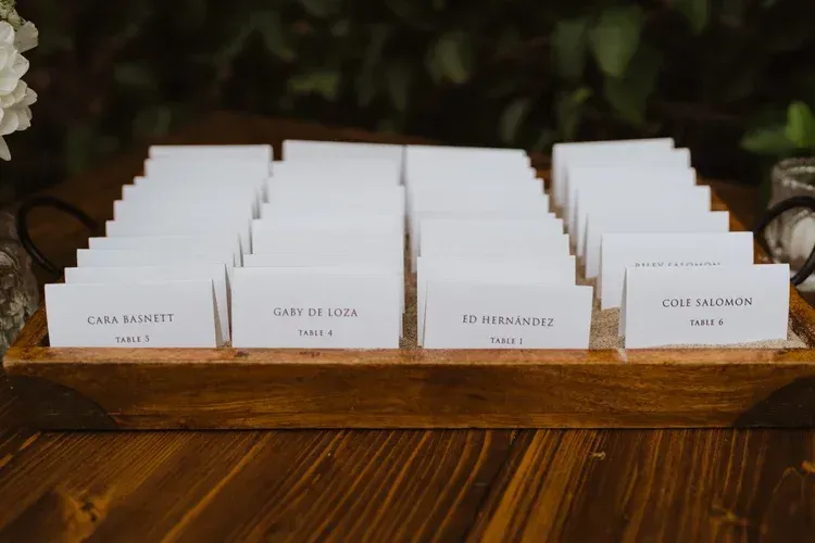 White name cards arranged in rows on a wooden table, with greenery blurred in the background