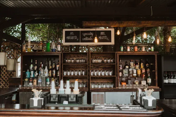Wooden bar with shelves of bottles and glassware, chalkboard menu, and coffee machines under warm lights