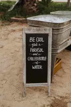 Chalkboard sign on sand offers free parasol, chilled bottle, and water beside stacked loungers.