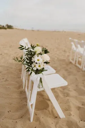 White chairs lined on a sandy beach, decorated with white flowers for an outdoor ceremony.
