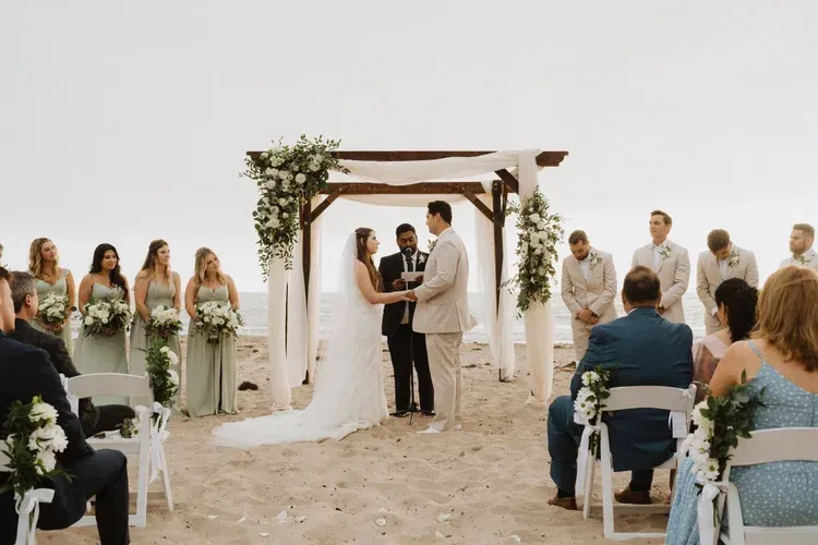Wedding ceremony under a floral arch on a beach, with bride, groom, officiant, and seated guests.