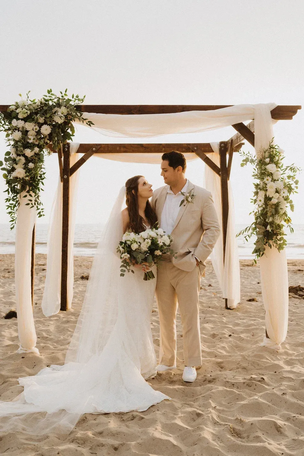 Bride and groom standing under a beach wedding arch, holding hands on sandy shore.