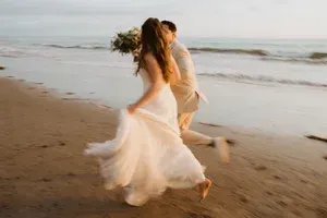 Couple running hand in hand on a beach, with the woman’s white dress flowing in the wind.