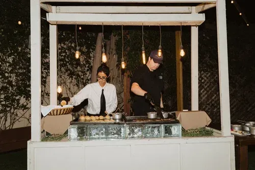 Two people cooking at an outdoor grill under a white pergola at night, with hanging lights and candles.