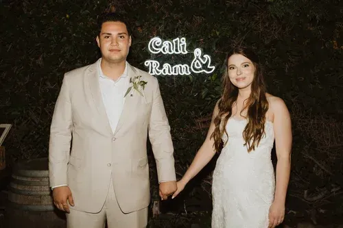 Couple in formal attire holding hands at a nighttime event with a leafy backdrop.