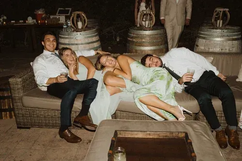 Four people reclining on a sofa at a rustic indoor gathering, with large barrels behind them.