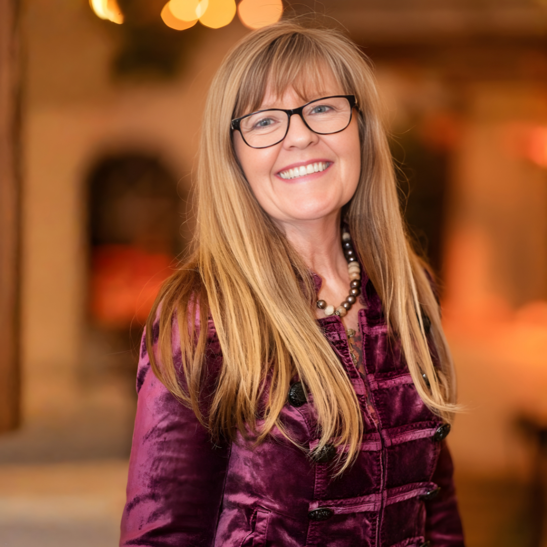 Smiling woman in glasses wearing a purple velvet jacket, standing indoors with a blurred warm-toned background