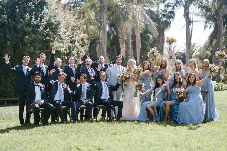 Large wedding party posing outdoors on a lawn with trees and flowers, bride in white, bridesmaids in blue.