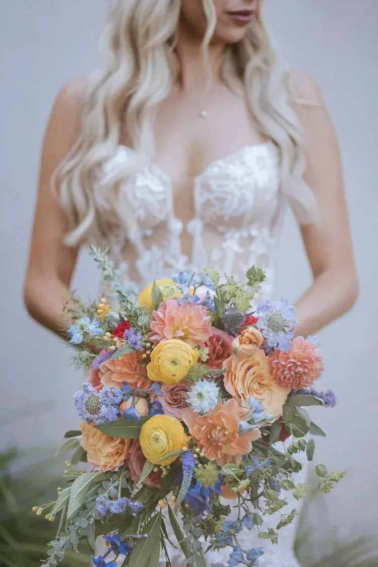Bride holding a colorful bouquet of yellow, peach, and lavender flowers in a white lace dress